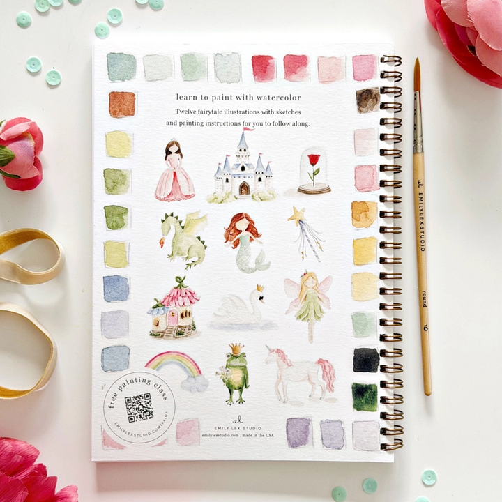 Watercolor art book with illustrations of fairies, castles, and other fantasy elements on a white surface with flowers and a pen.